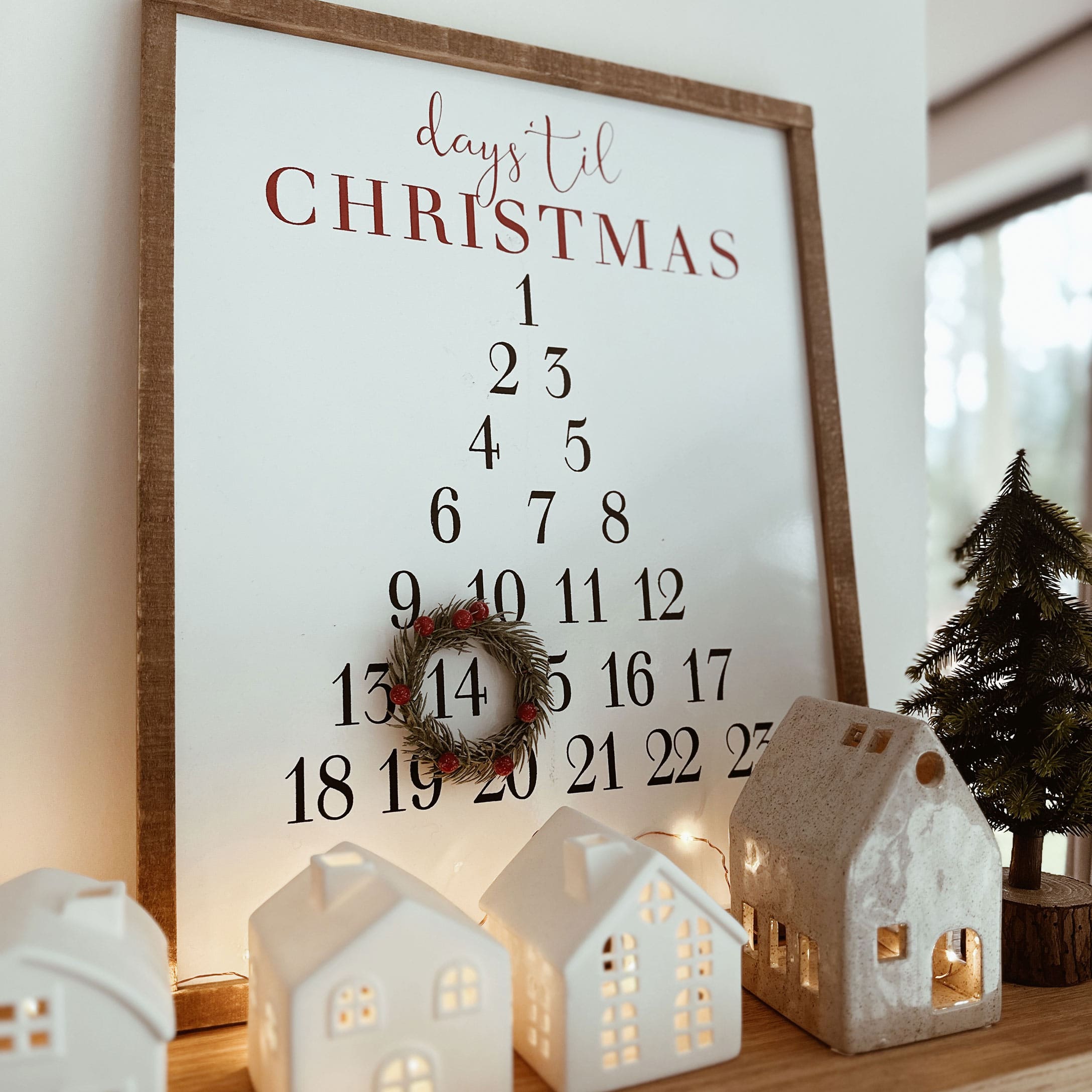 Holzrahmen mit Adventskalender und dekorativem Kranz, Keramikhäuser und Tannenbaum auf Tisch