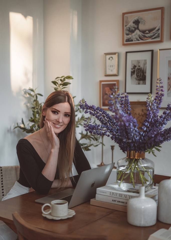 Frau am Holztisch mit Laptop und Glasvase mit violetten Blumen in hellem Raum