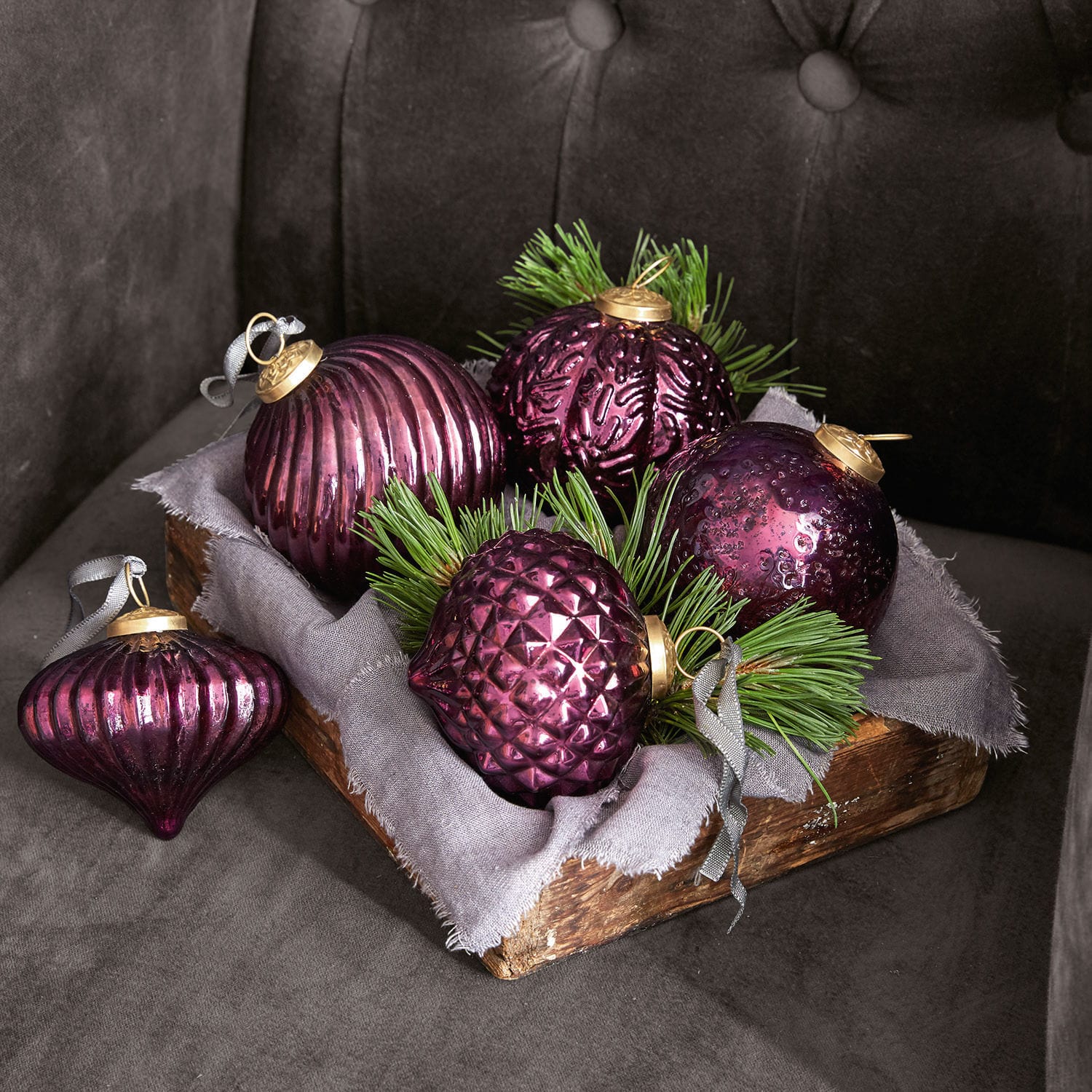 Weihnachtsschmuck 5er Set Sambre Set aus violetten Glas-Weihnachtskugeln mit grünen Zweigen in Holzkiste auf grauem Stoff