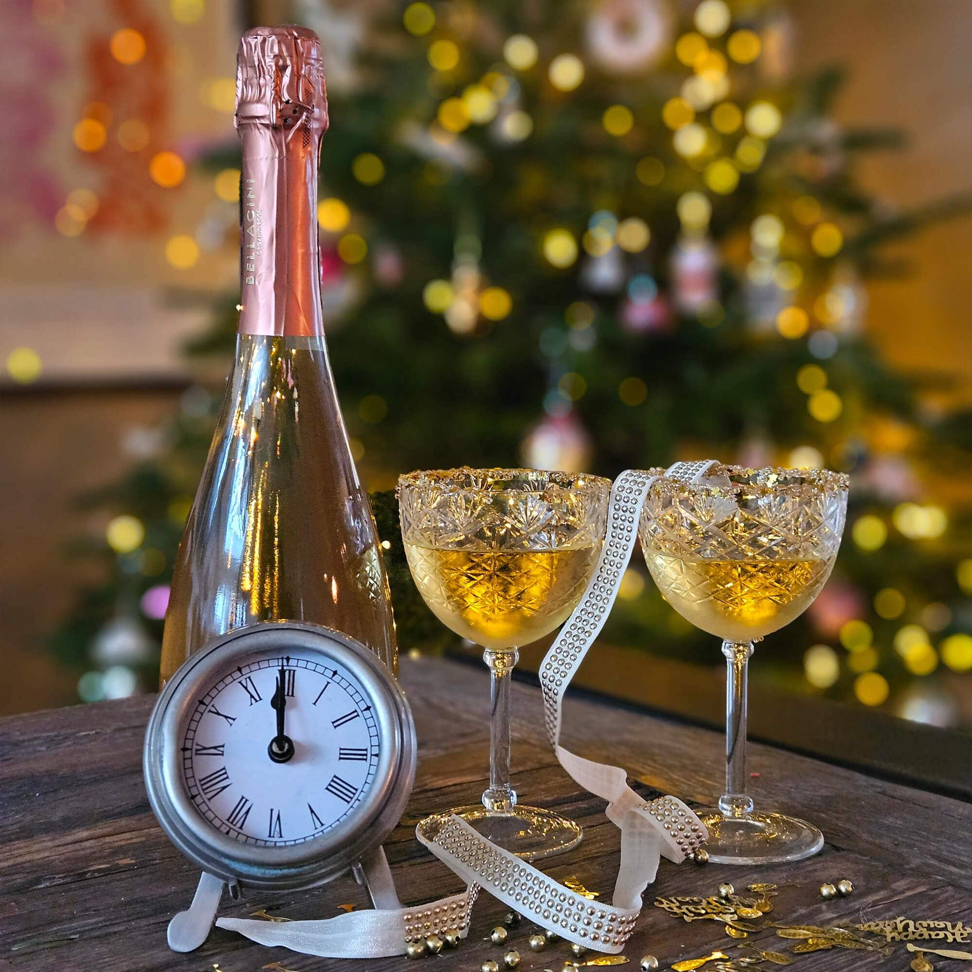 Flasche Champagner mit rosafarbener Folie, zwei Kristallgläser mit goldenem Getränk und Uhr auf Holztisch