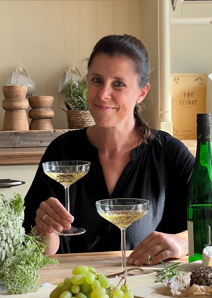 Frau in schwarzer Bluse hält Weißweinglas am Tisch mit Trauben und Käse