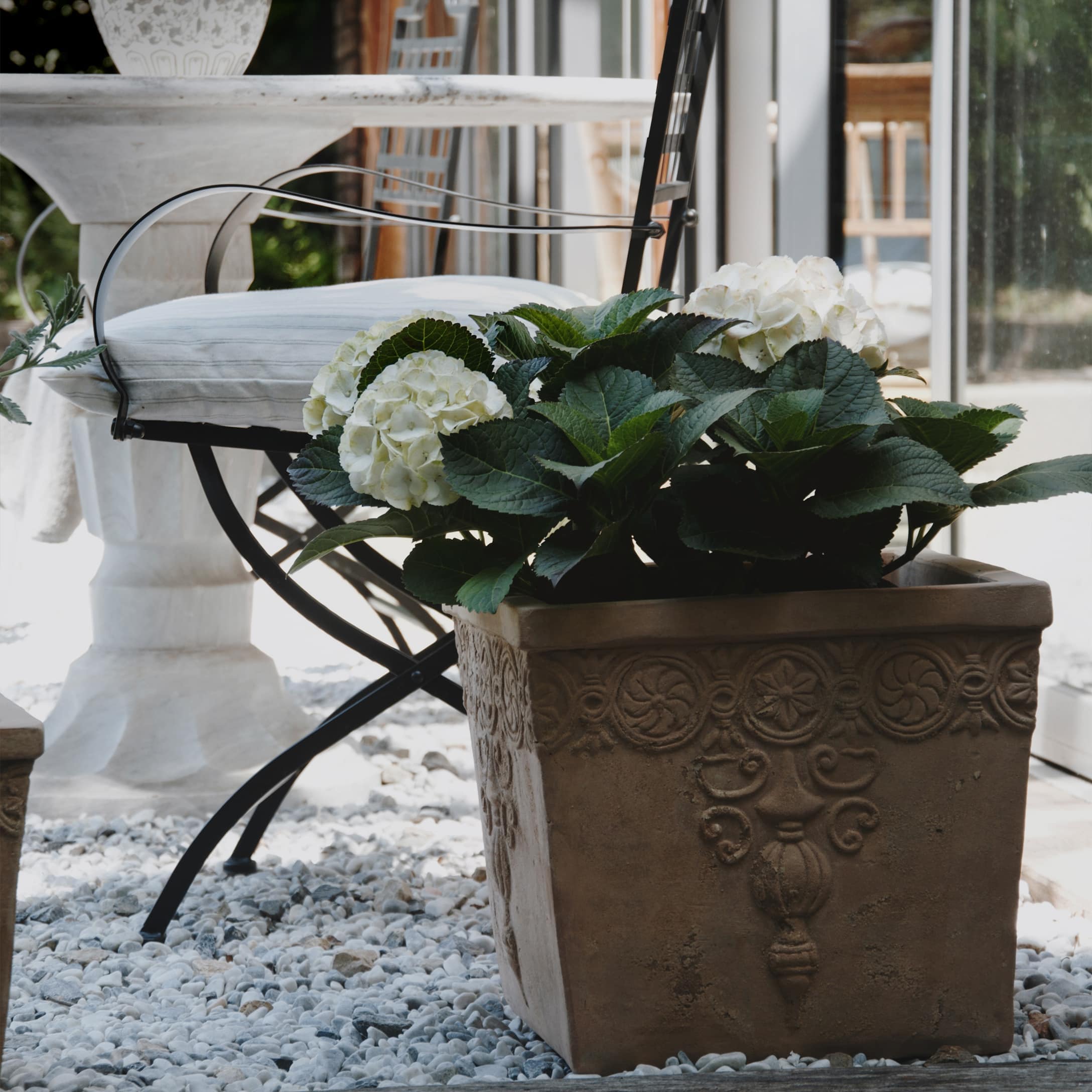Braune Keramik-Pflanzkübel mit weißen Hortensien auf Kieselsteinboden neben Metallstuhl