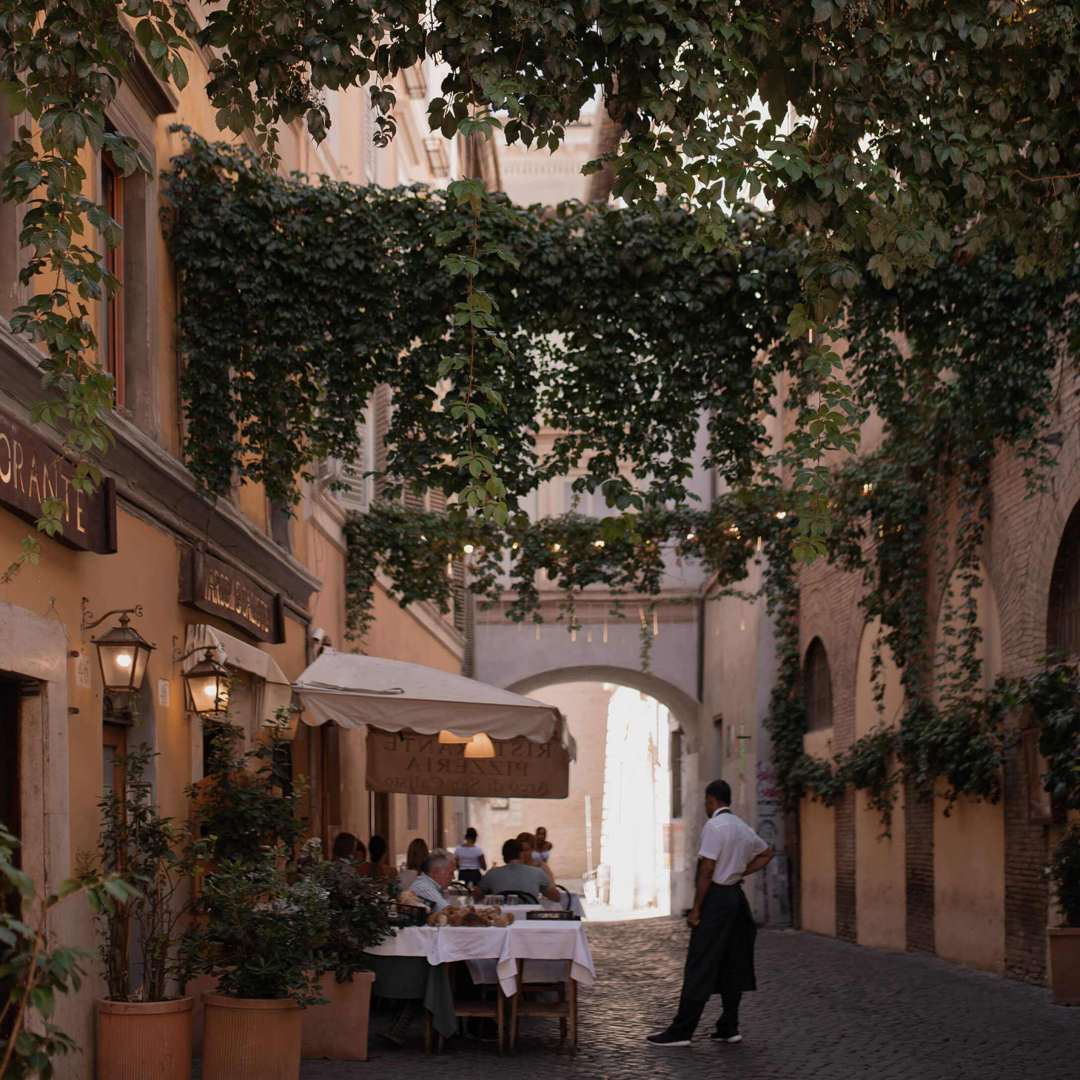 Pflastergasse mit Restaurantterrasse und grünen Kletterpflanzen, gemütliche Atmosphäre