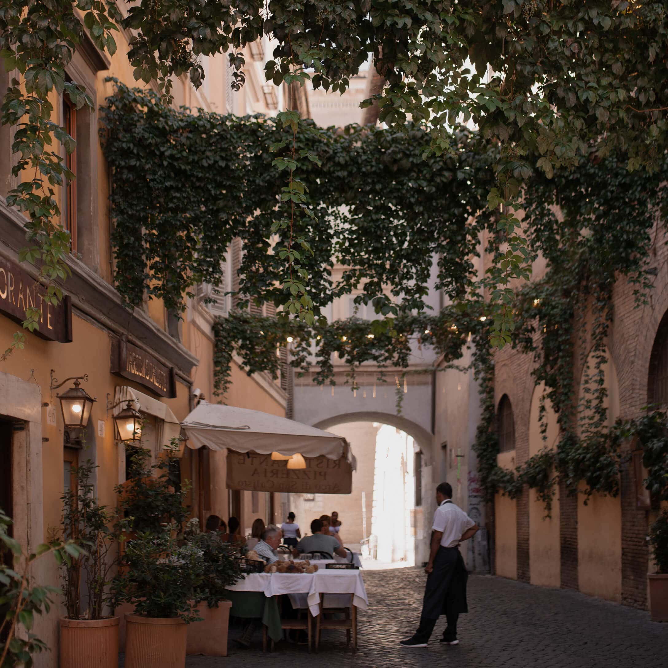 Pflastergasse mit Restaurantterrasse und grünen Kletterpflanzen, gemütliche Atmosphäre