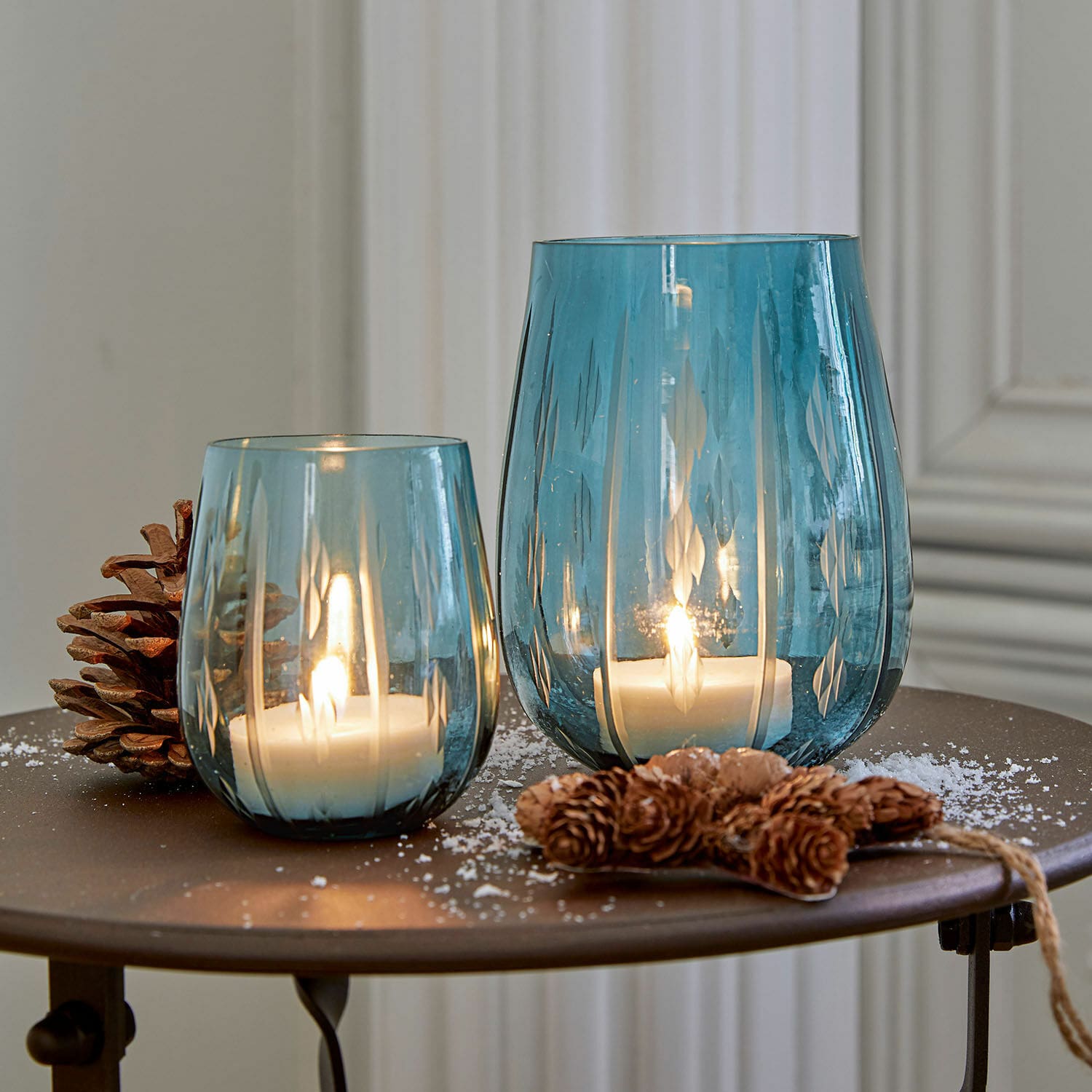 Windlicht 2er Set Danisar Zwei blaue Glaswindlichter mit Muster, weißen Kerzen und Kiefernzapfen-Dekoration auf Tisch