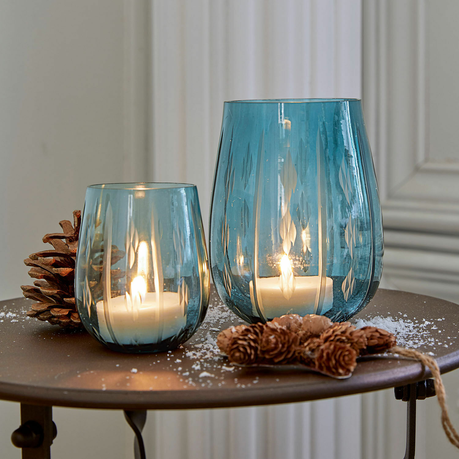 Windlicht 2er Set Danisar Zwei blaue Glaswindlichter mit Muster, weißen Kerzen und Kiefernzapfen-Dekoration auf Tisch