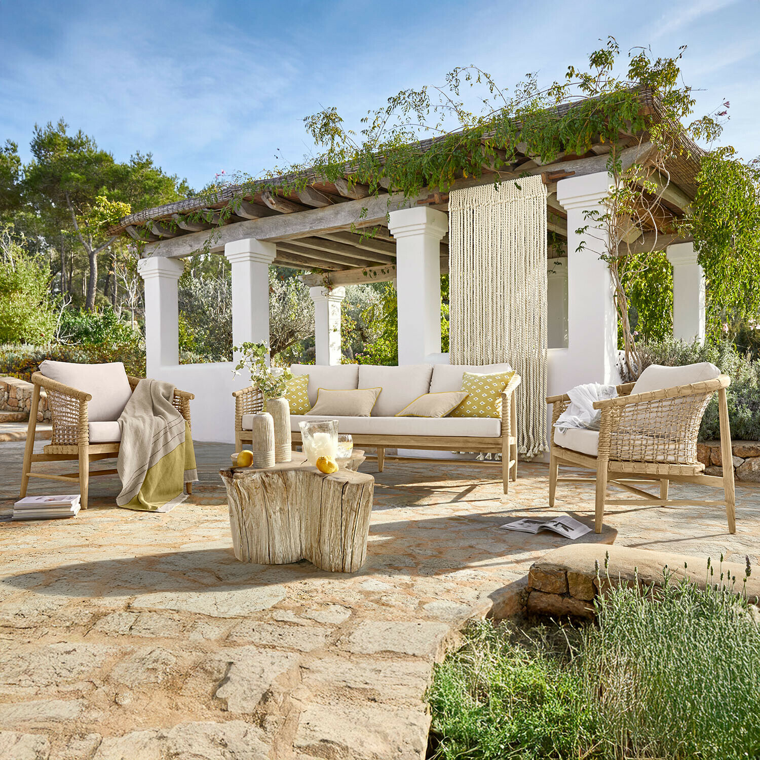 Sitzgruppe Roselva Gartenmöbel-Set aus hellem Holz mit beigen Polstern auf Steinpatio und natürlichem Holztisch