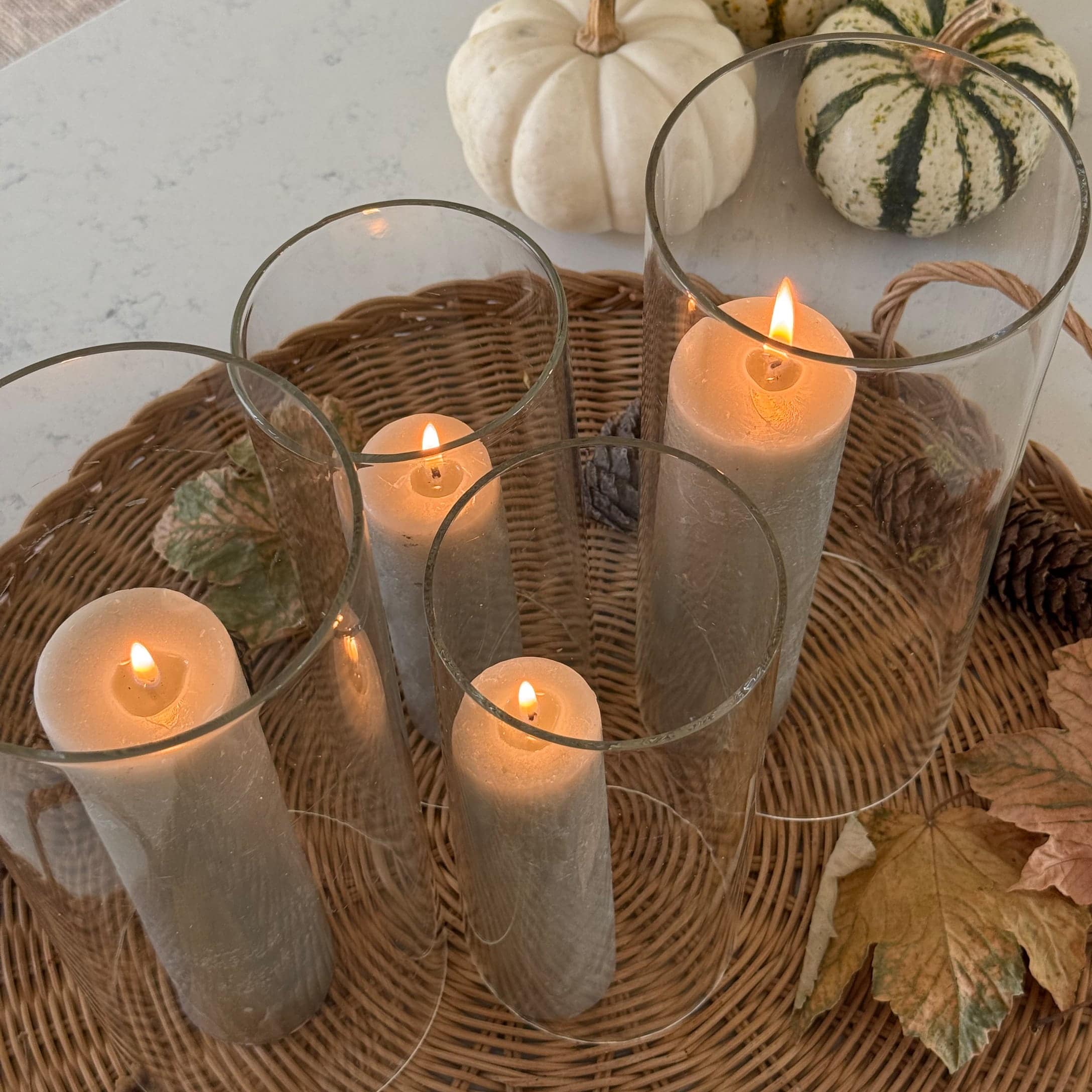 Vier weiße Kerzen in Glaszylindern auf einem Korbtablett mit Herbstblättern und Kürbissen