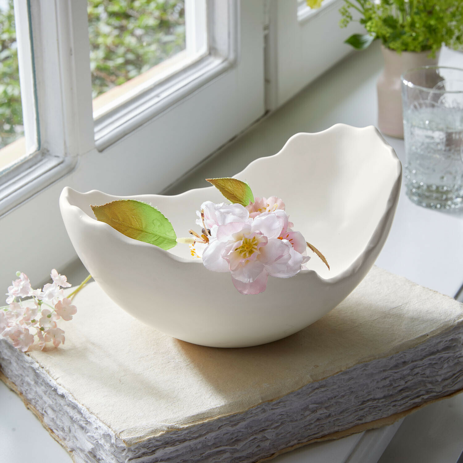 Weiße Keramikschale mit wellenförmigem Rand und zarter Blüte auf Buch am Fensterbrett