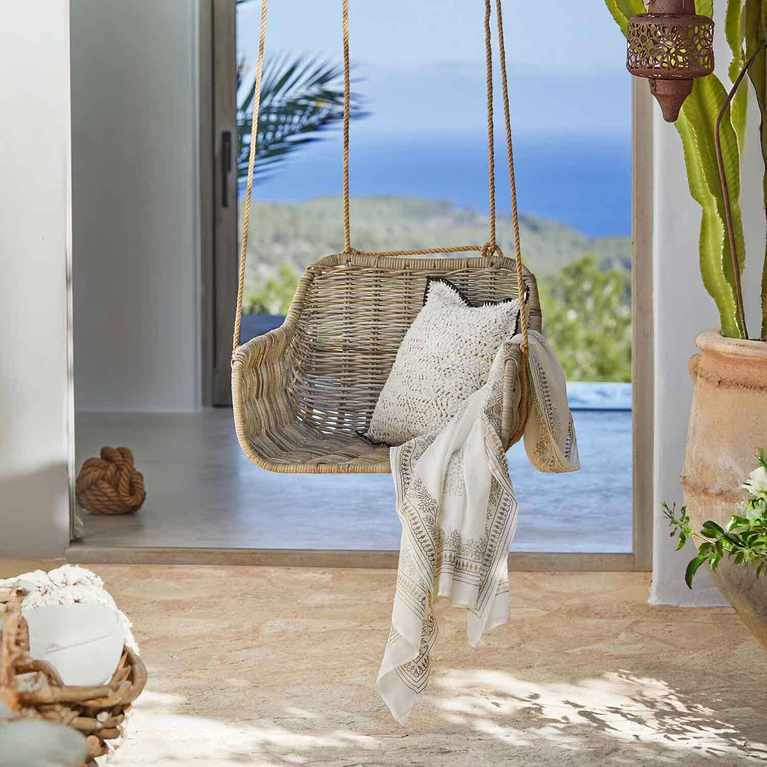 Rattanschaukel in Naturfarbe mit weißem Kissen und dekorativem Tuch in hellem Raum