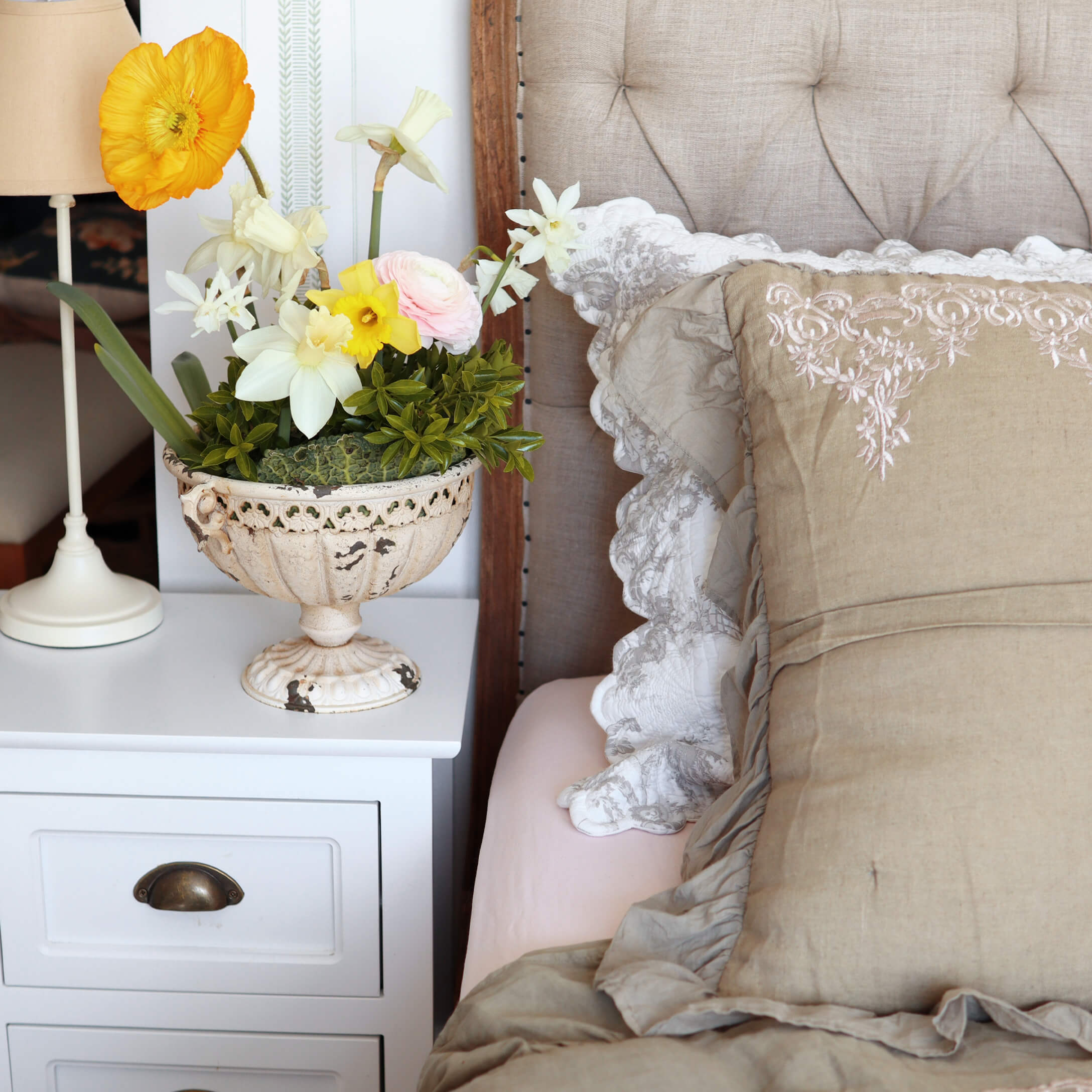 Dekorative weiße Keramikvase mit bunten Blumen auf weißem Nachttisch neben dem Bett