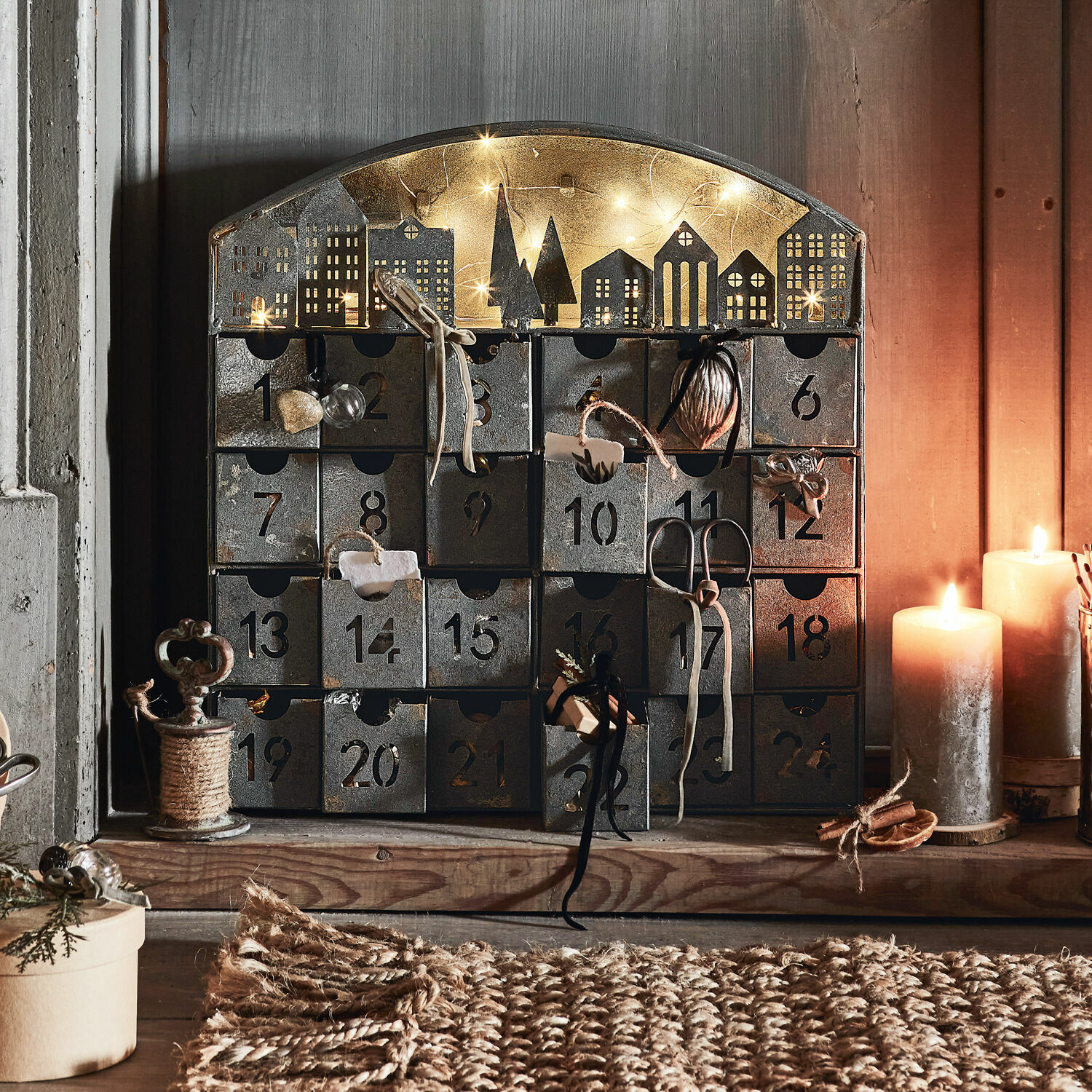 Adventskalender Léran Metall-Adventskalender mit 24 nummerierten Schubladen und beleuchteter Häusersilhouette, dekoriert mit Kerzen
