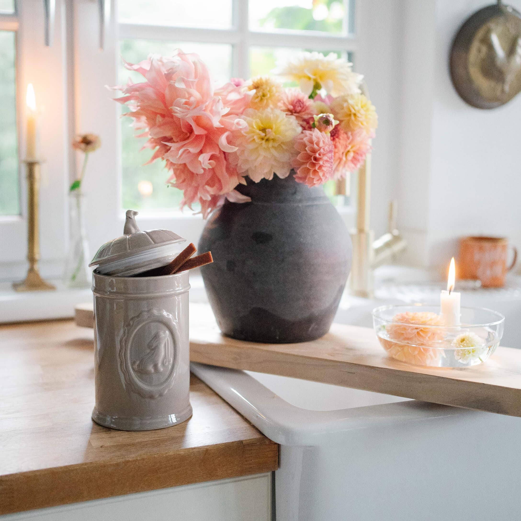 Grauer Keramikbehälter mit Deckel auf Holzarbeitsplatte neben schwarzer Vase mit rosa-gelben Blumen
