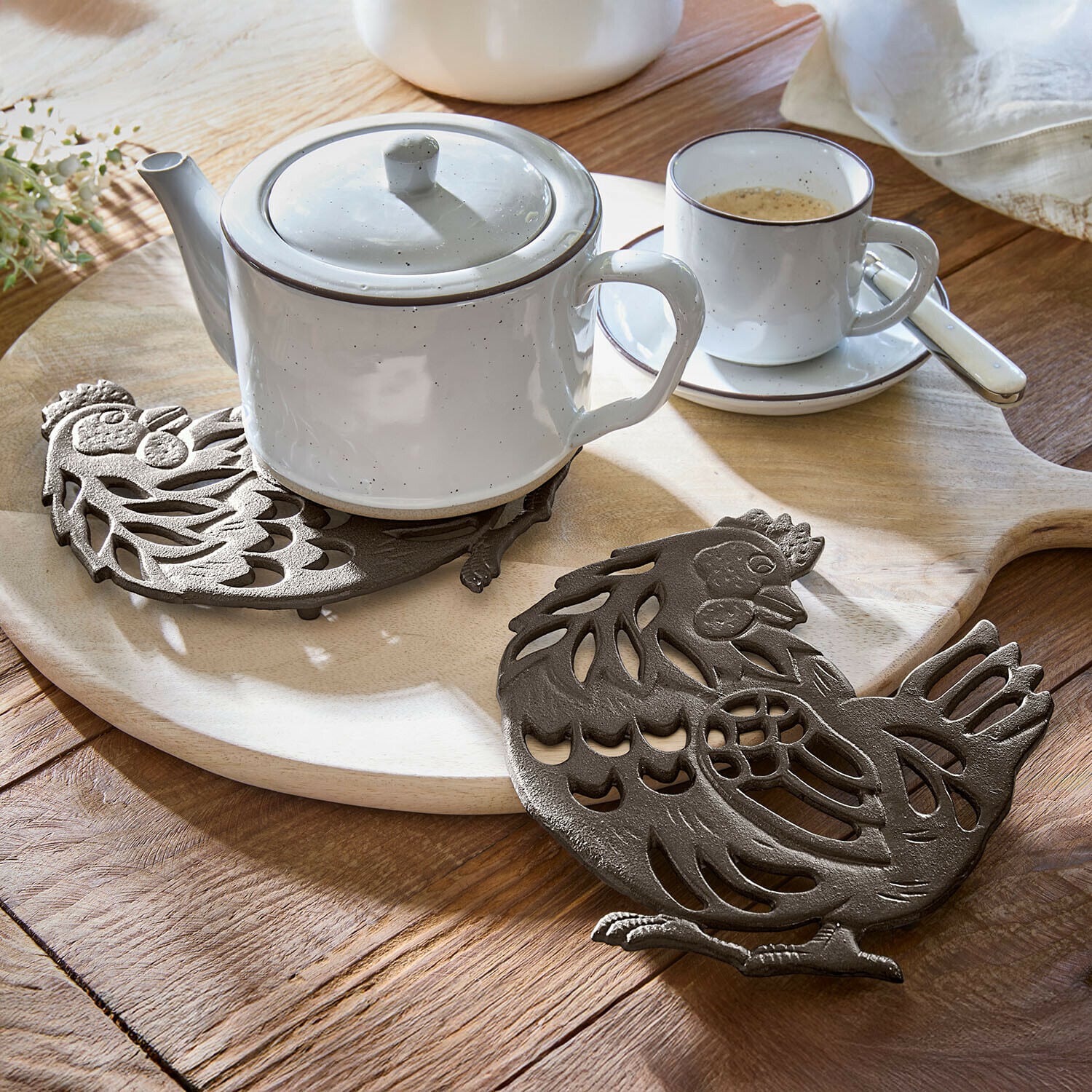Brauner gusseiserner Topfuntersetzer in Hühnerform auf Holztisch mit weißer Teekanne und Tasse