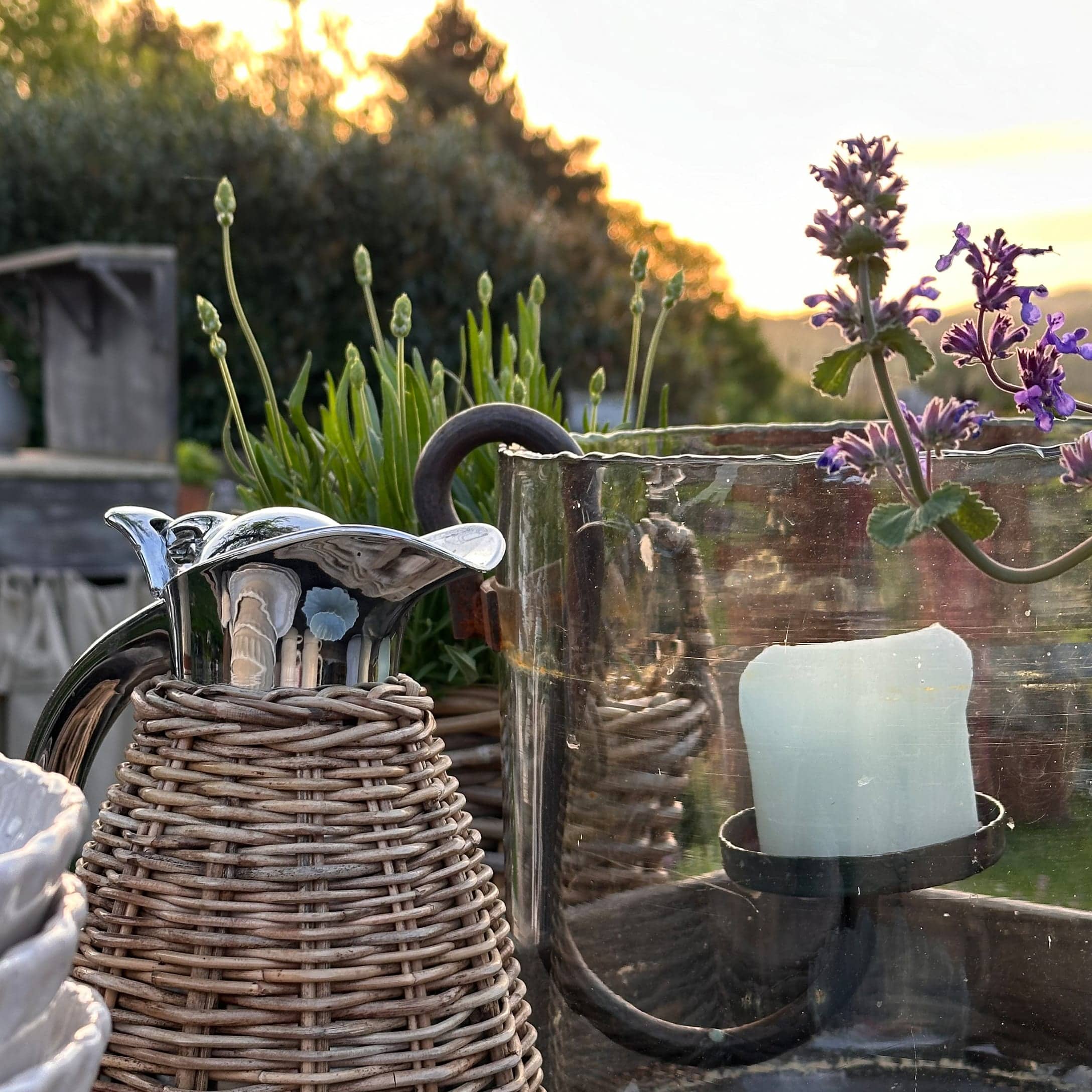 Metallkrug mit Korbgeflecht und Glas-Kerzenhalter in sommerlichem Garten
