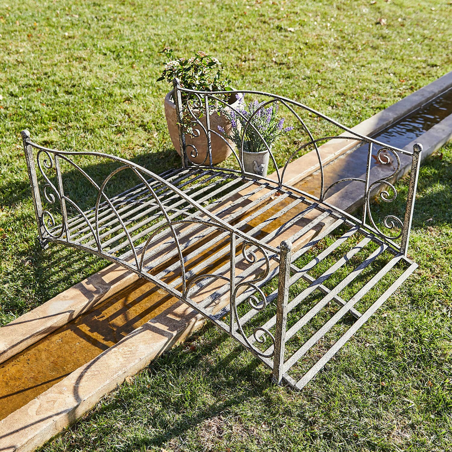 Deko-Brücke Escàs Dekorativer Gartenbrücke aus Metall in antik-silberner Optik über einem Wasserlauf auf Rasen