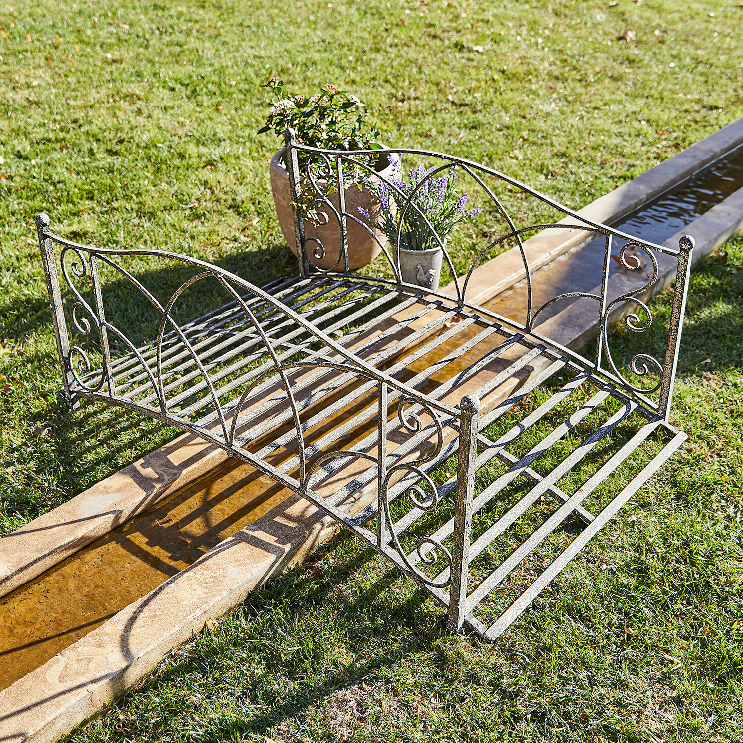 Deko-Brücke Escàs Dekorativer Gartenbrücke aus Metall in antik-silberner Optik über einem Wasserlauf auf Rasen