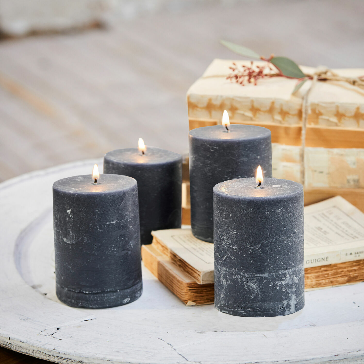 Kerze 4er Set Virrans Vier schwarze Wachskerzen auf weißem Holztisch mit Büchern und Geschenk im Hintergrund