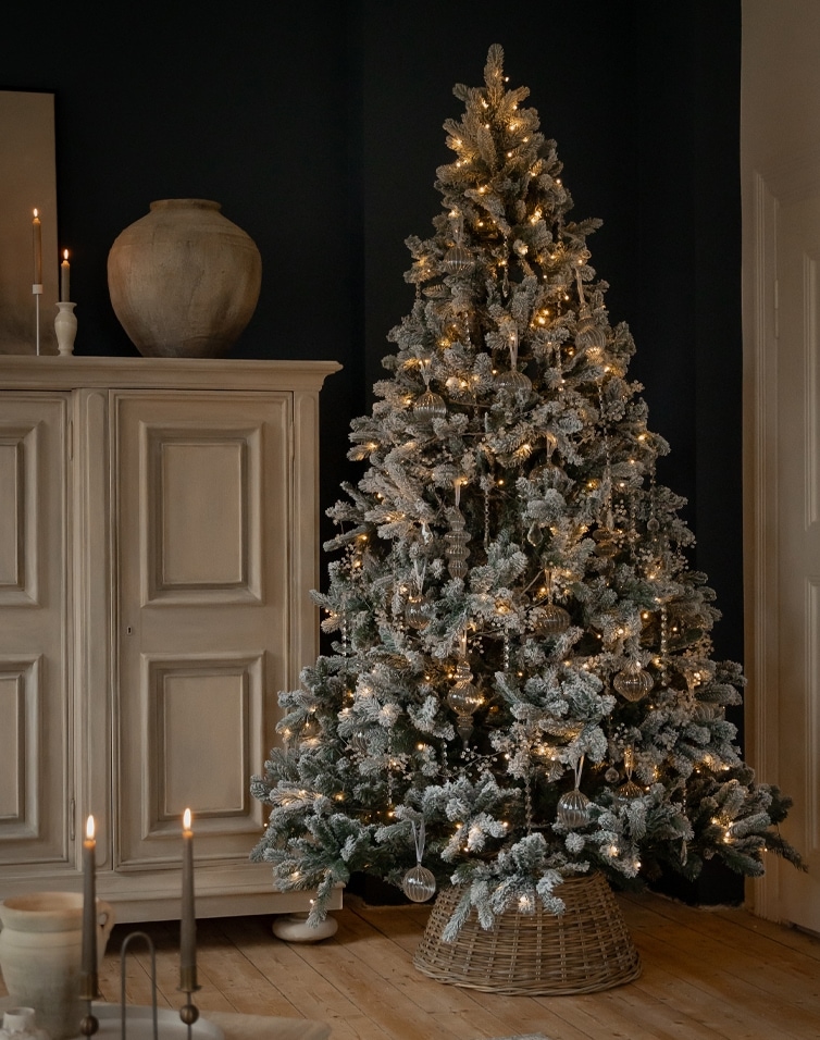 Künstlicher, schneebedeckter Weihnachtsbaum mit warmen Lichtern und Glasornamenten im Korbständer