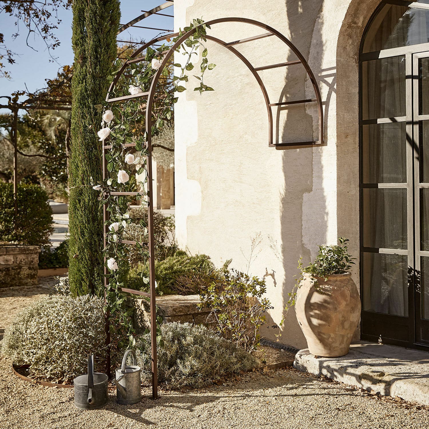 Rosenbogen Auburn Braune Metall-Gartenpergola mit Bogen, weißen Rosen und Gießkannen auf Kiesboden