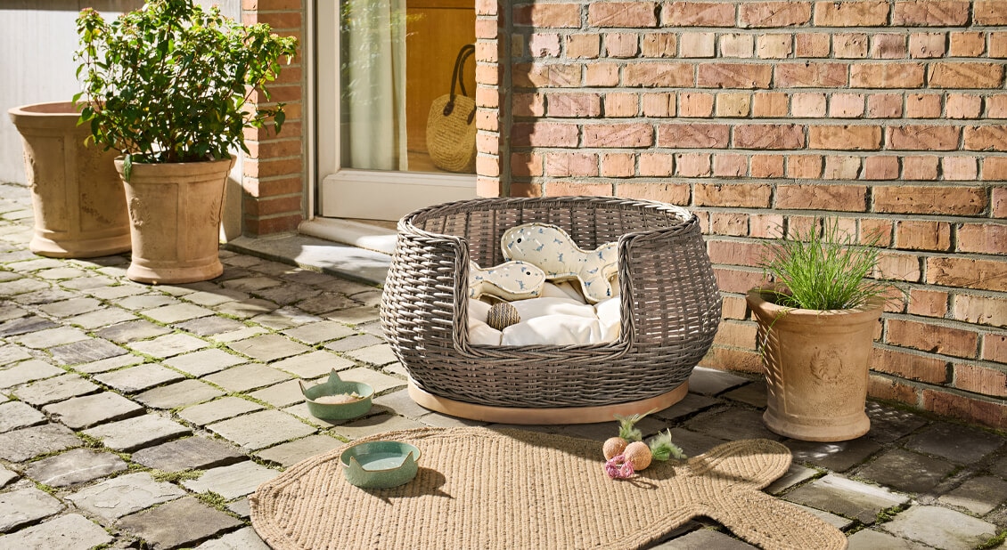 Rundes Katzenbett aus grauem Rattan mit Kissen und Spielzeug auf gepflasterter Terrasse