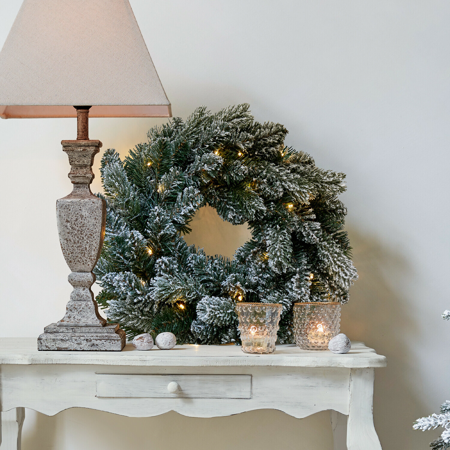 LED-Kranz Regin Schneebedeckter Weihnachtskranz mit grünen Zweigen und Lichterkette auf weißem Tisch mit Lampe
