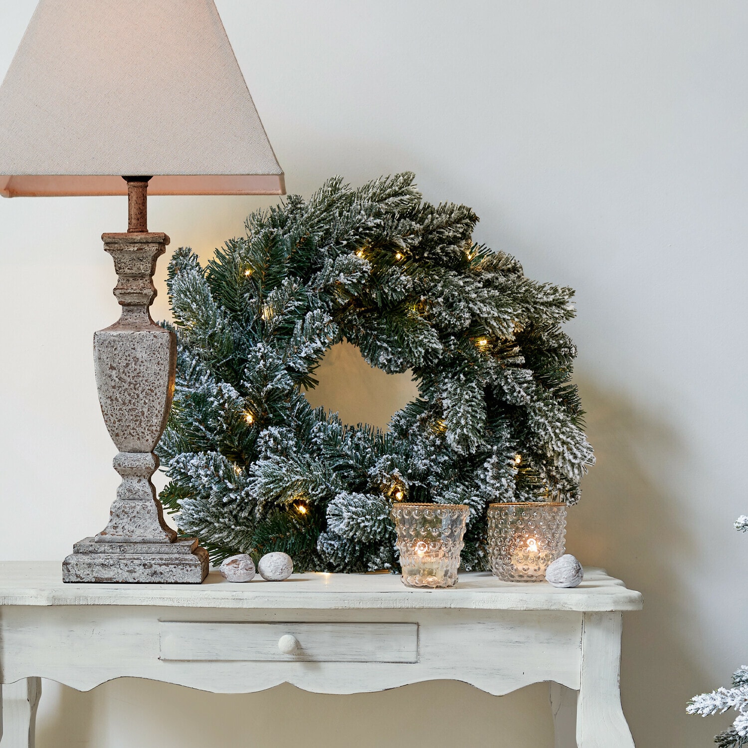 LED-Kranz Regin Schneebedeckter Weihnachtskranz mit grünen Zweigen und Lichterkette auf weißem Tisch mit Lampe