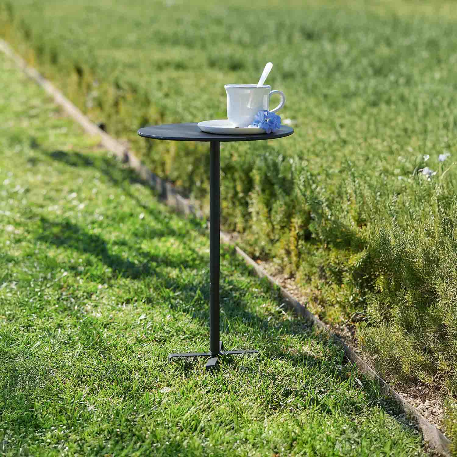 Kleiner schwarzer Metall-Gartentisch mit weißer Tasse und blauer Blume auf Gras