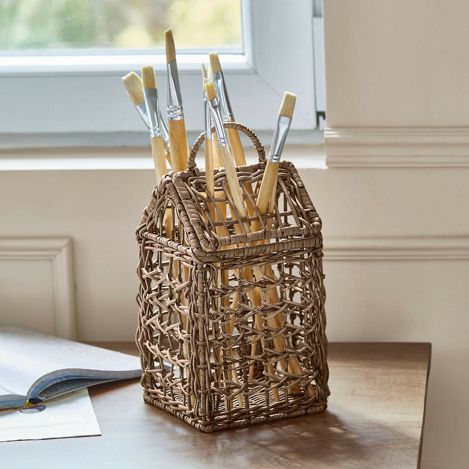 Brauner geflochtener Aufbewahrungskorb in Hausform mit Malpinseln auf Holztisch