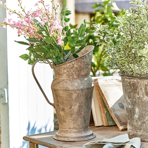 Metallkrug im Vintage-Stil mit grünen und rosa Blumen auf Gartentisch bei Tageslicht