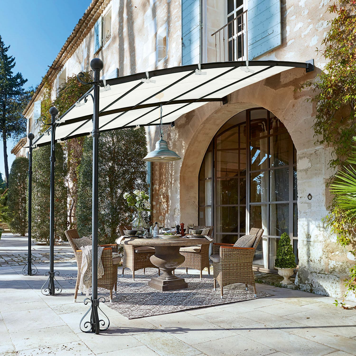 Pergola Kattles Gartenmöbel-Set aus Rattan unter weißem Vordach auf steinerner Terrasse vor einem Steinhaus