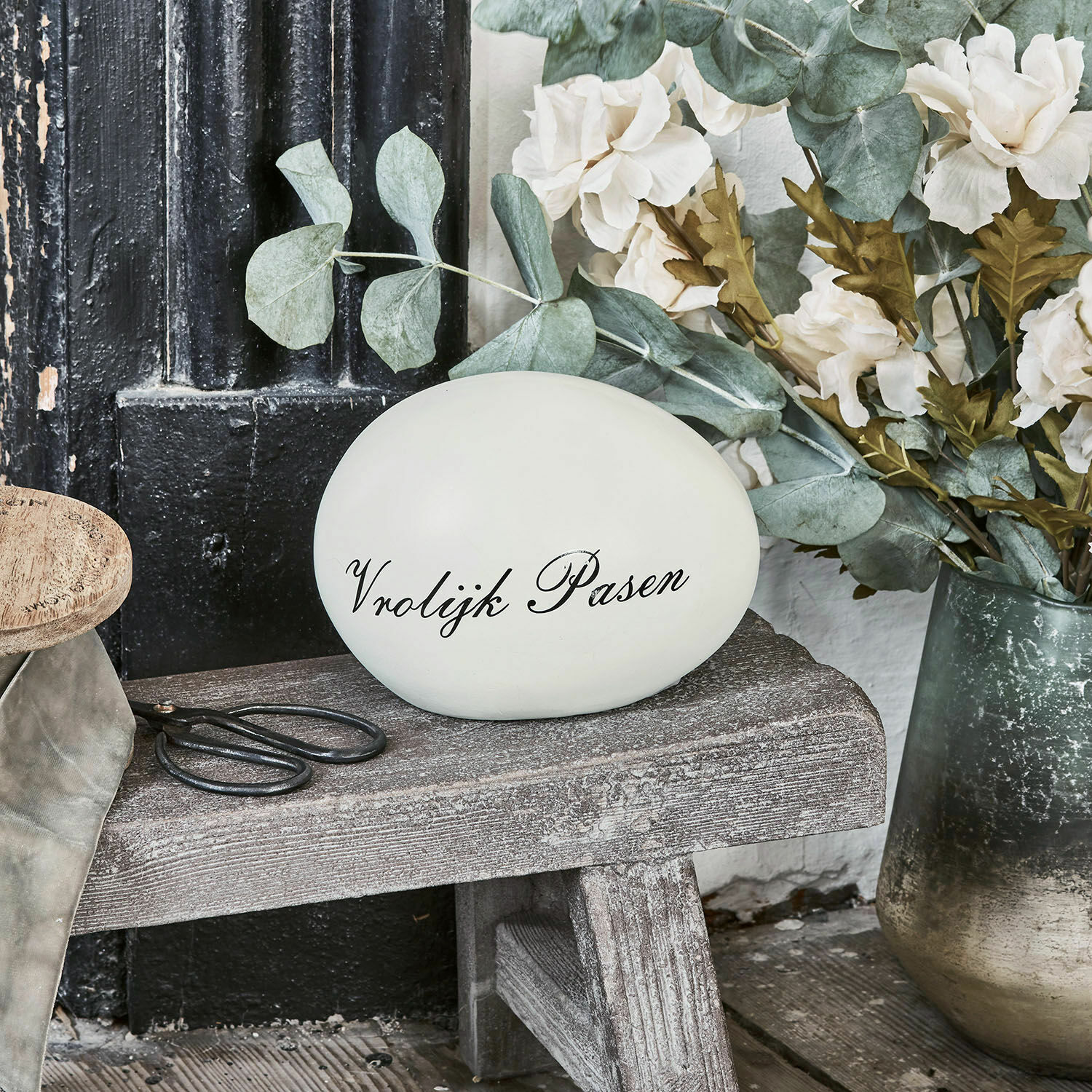 Deko-Ei VROLIJK PASEN Dekoratives weißes Ei mit Schriftzug Vrolijk Pasen auf Holzbank neben Blumen und Schere