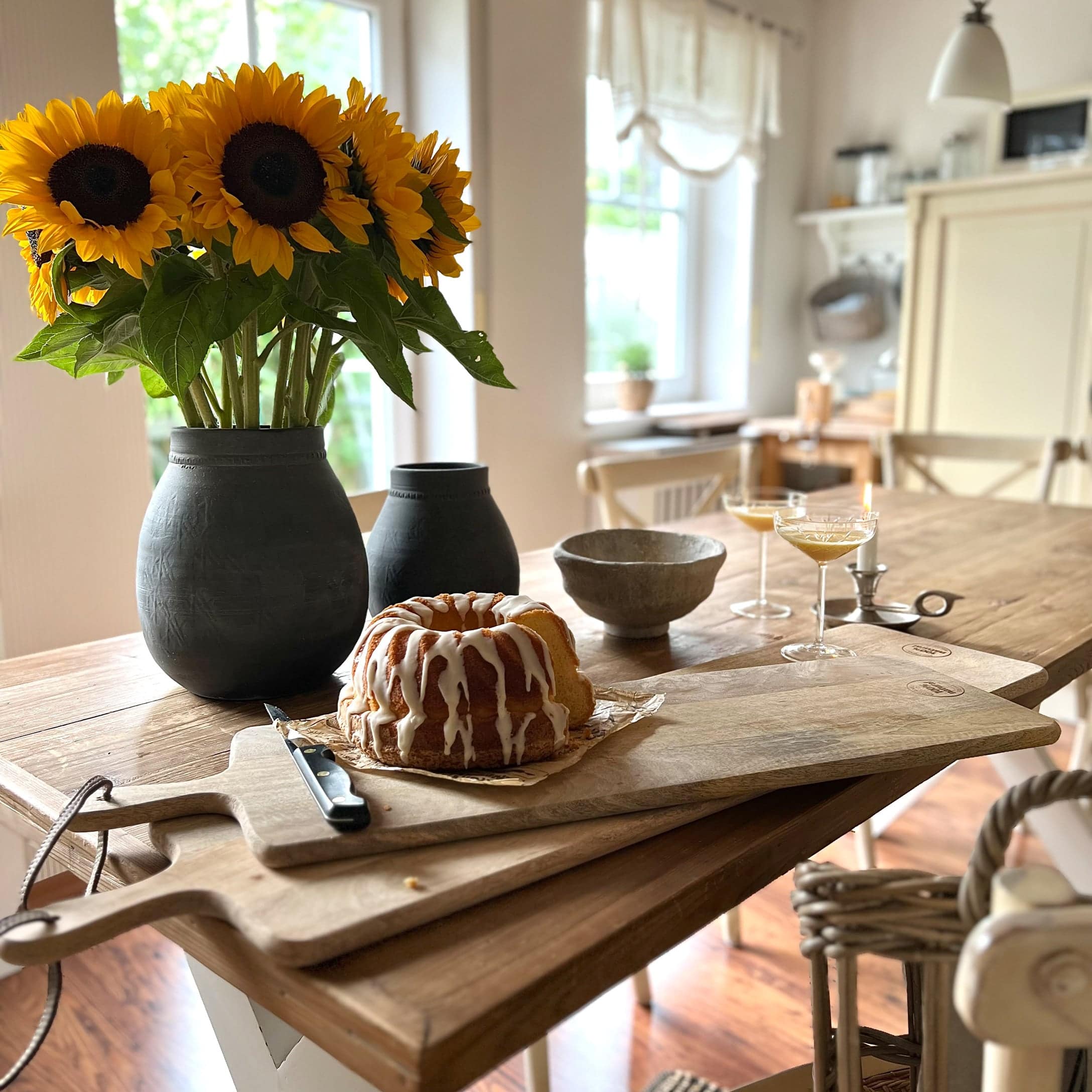 Holz-Schneidebrett mit Kuchen und schwarzem Vase mit gelben Sonnenblumen auf Holztisch in Küche
