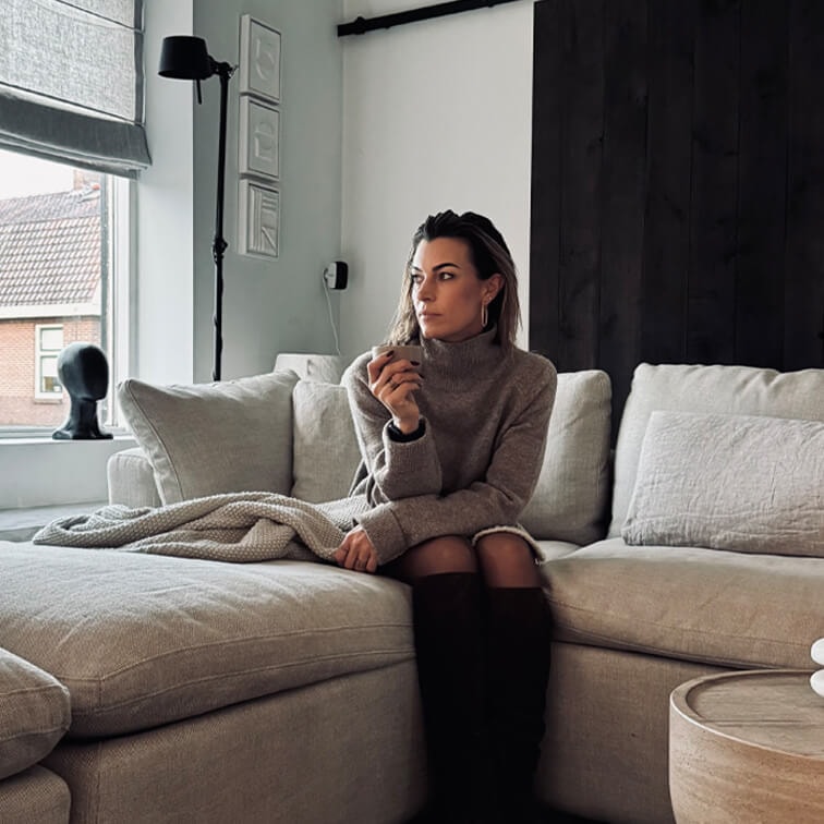Frau im grauen Pullover sitzt auf beigem Sofa in modernem Wohnzimmer mit großem Fenster
