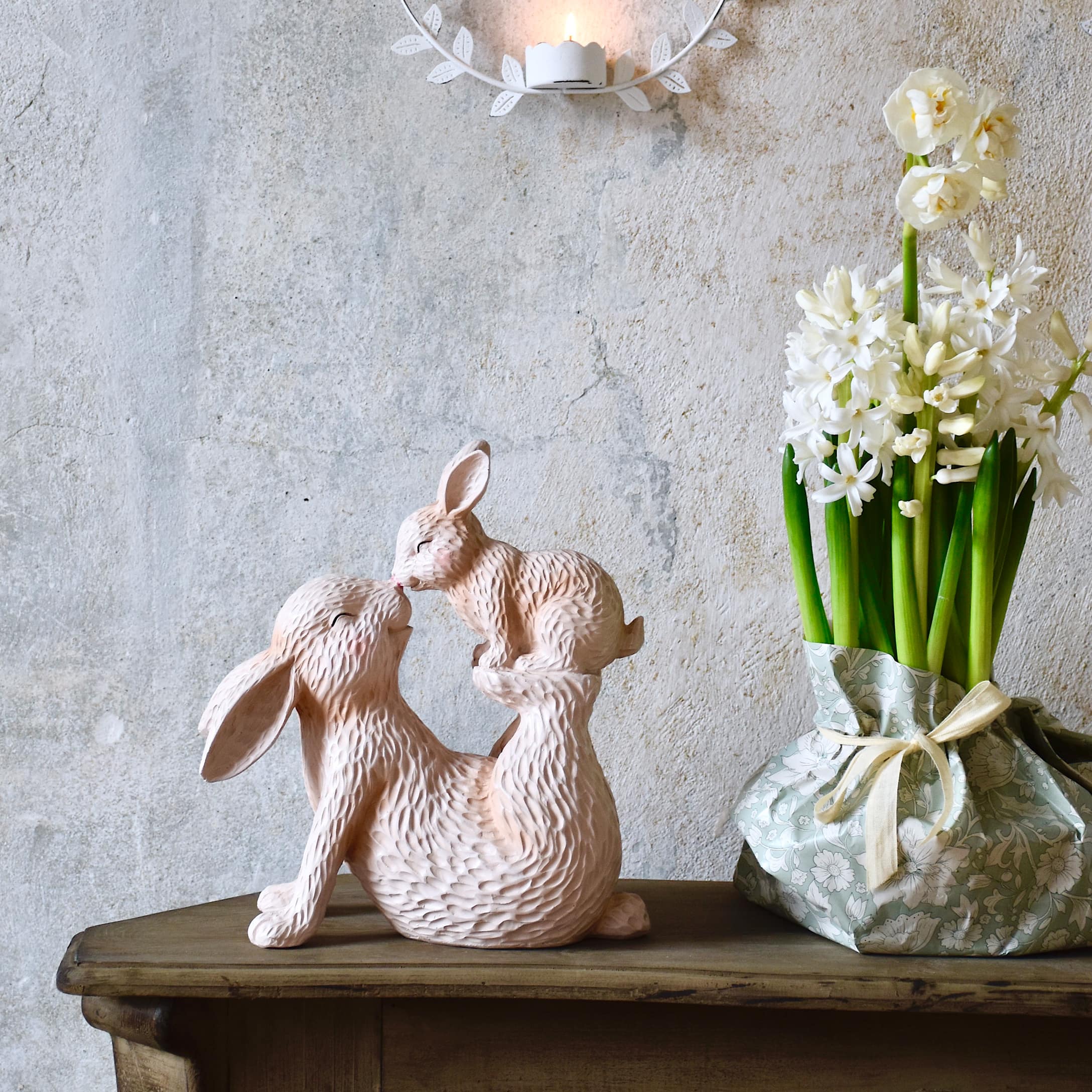 Rosa Holzskulptur mit zwei Hasen auf einem Holztisch neben Blumen in pastellfarbenem Übertopf