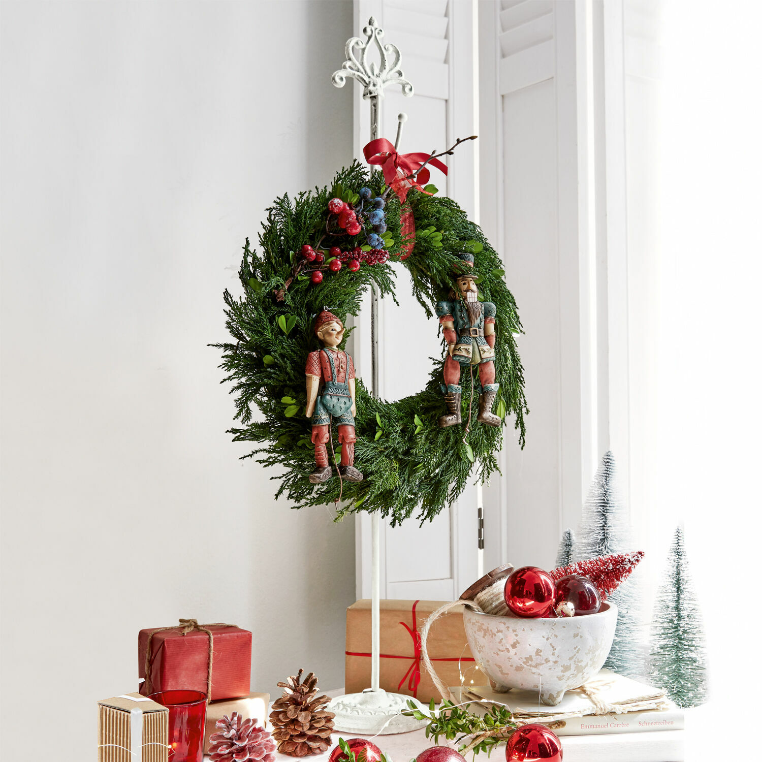 Weihnachtskranz aus grünen Zweigen mit roten und blauen Beeren und Holzfiguren auf weißem Ständer