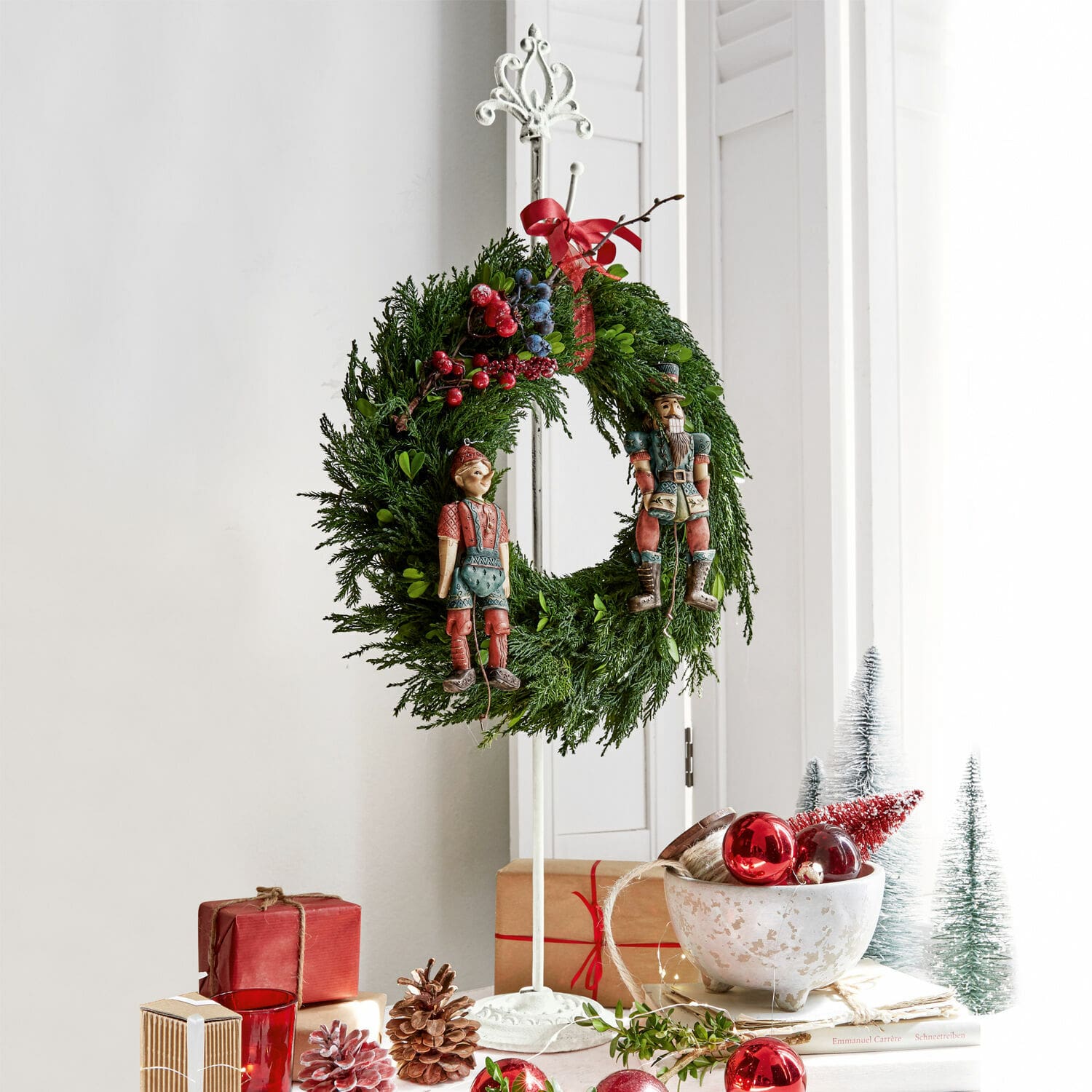 Kranzständer Irulan Weihnachtskranz aus grünen Zweigen mit roten und blauen Beeren und Holzfiguren auf weißem Ständer