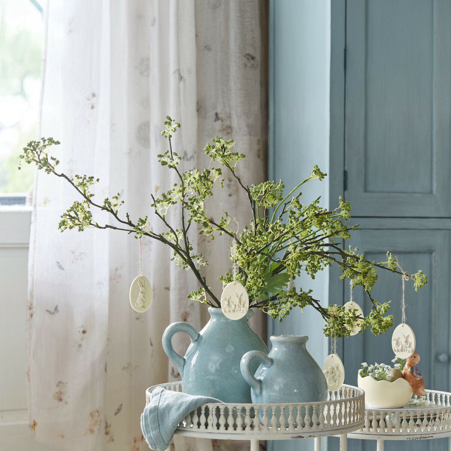 Blaue Keramikkrüge mit grünen Zweigen und Osterdekorationen auf weißem Metalltisch