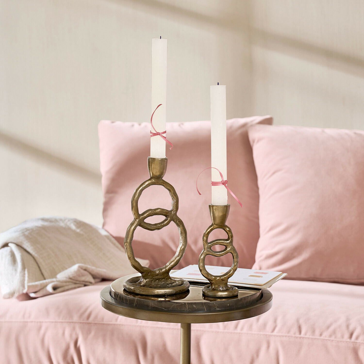 Kerzenständer 2er Set Corvane Set aus zwei messingfarbenen Kerzenhaltern mit weißen Kerzen auf einem Tisch neben einem rosa Sofa