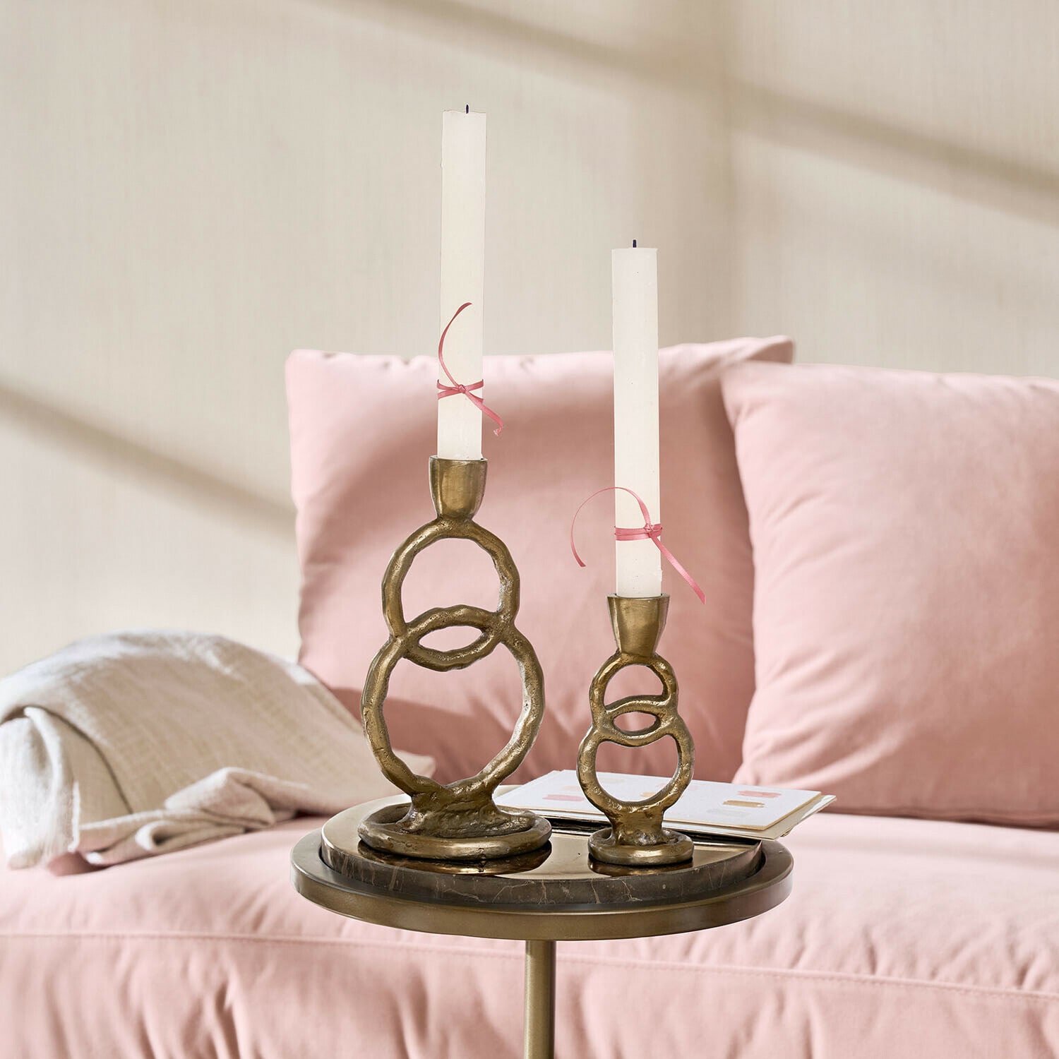 Kerzenständer 2er Set Corvane Set aus zwei messingfarbenen Kerzenhaltern mit weißen Kerzen auf einem Tisch neben einem rosa Sofa