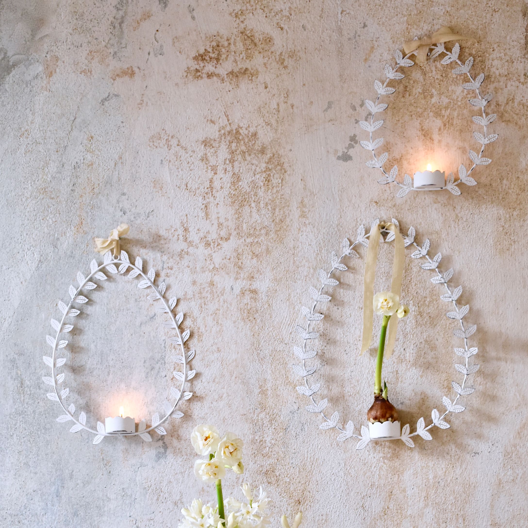 Weiße Metallrahmen in Ei-Form mit Blättern, Kerzen und Blumen an rustikaler Wand dekoriert
