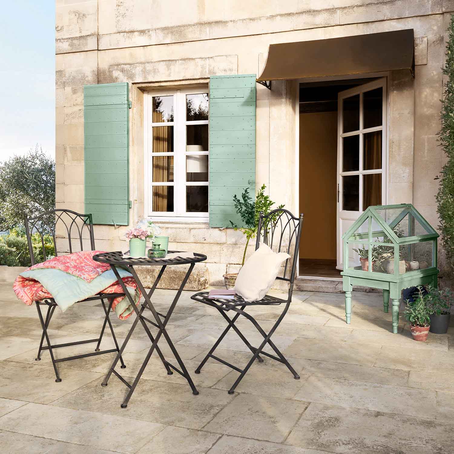Metall-Gartenmöbel mit Tisch und Stühlen auf Terrasse, grüne Mini-Gewächshaus und pastellfarbene Accessoires