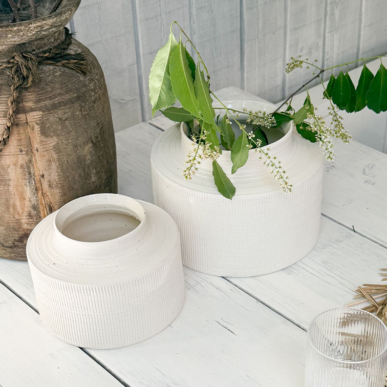 Weiße, matte Keramikvasen auf Holztisch mit grünen Blättern und kleinen weißen Blüten