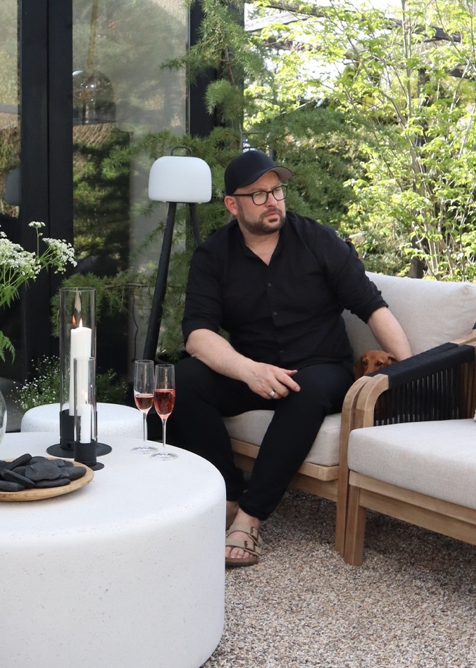 Mann in schwarzer Kleidung sitzt auf beigem Sofa im Garten mit Lampe und Kerzen auf weißem Tisch
