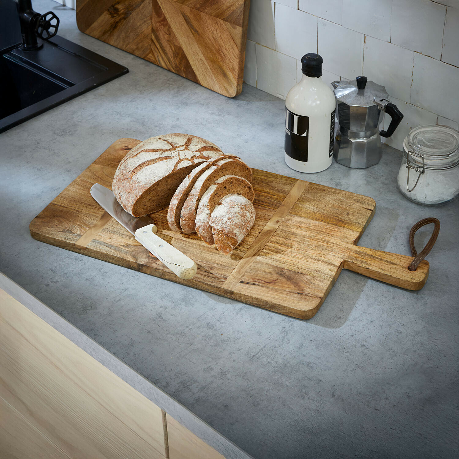 Schneidebrett aus Mangoholz mit Griff, darauf geschnittenes Brot und Messer auf Betonarbeitsplatte