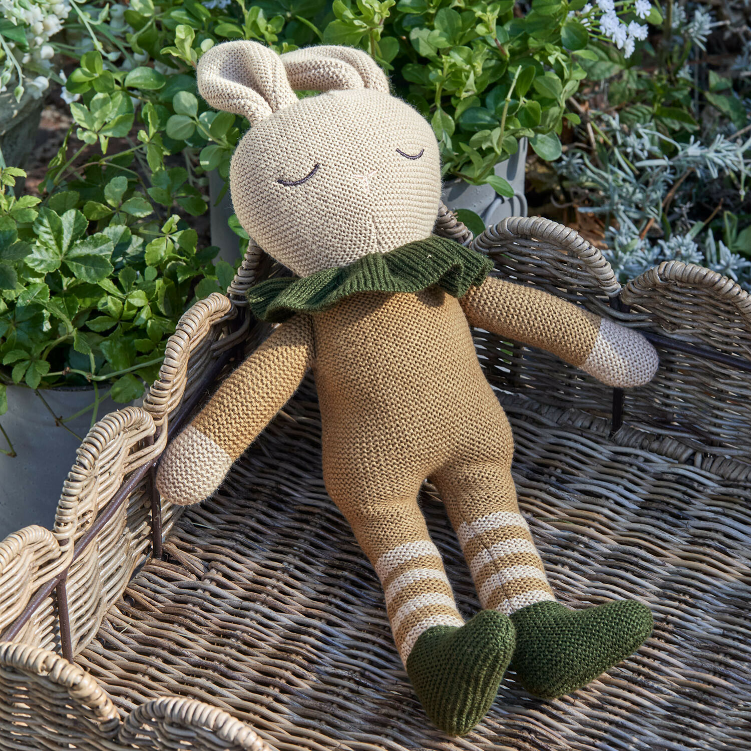 Beige gestricktes Hasenstofftier mit grünen Schuhen und Kragen auf einem Korb mit Pflanzen im Hintergrund