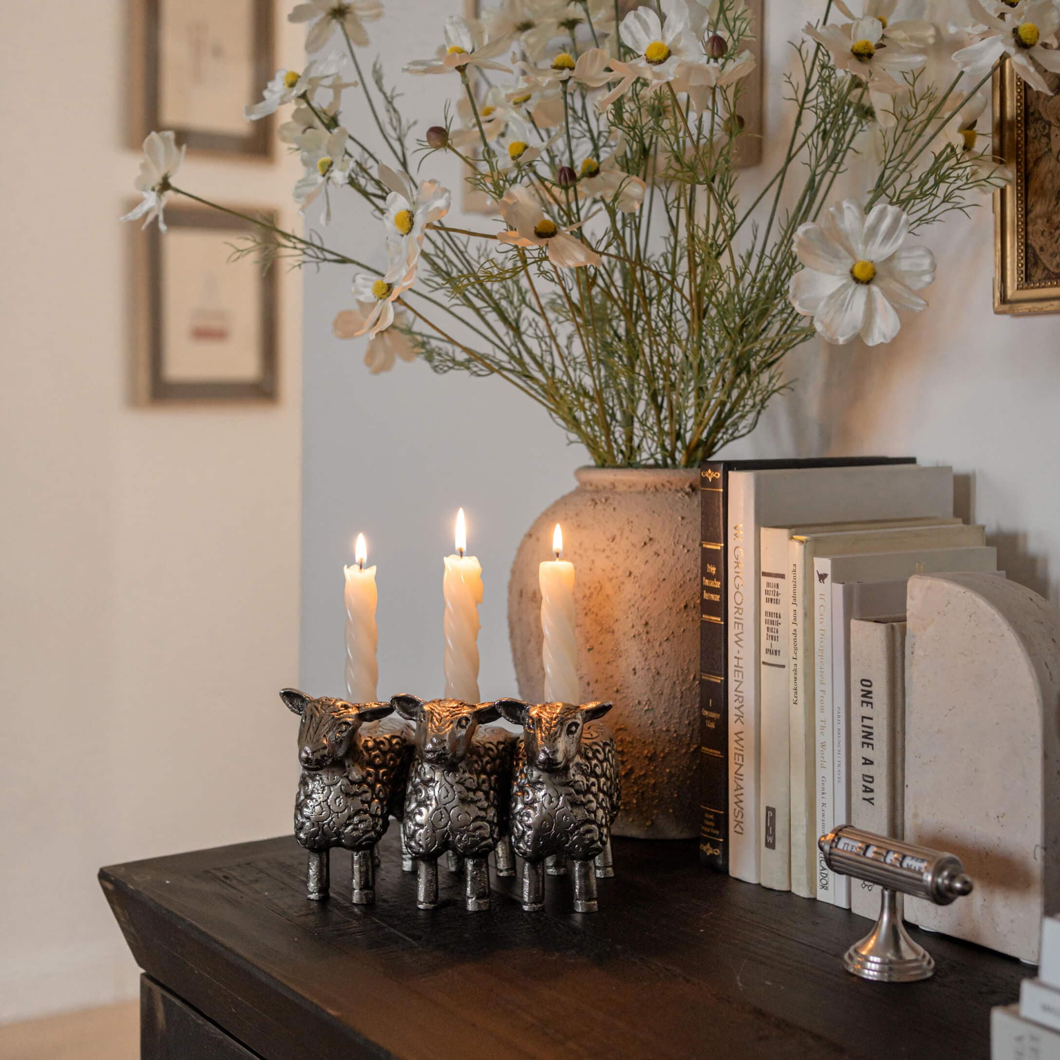 Metall-Kerzenhalter in Form von drei Schafen mit weißen Kerzen auf dunklem Holztisch neben Büchern