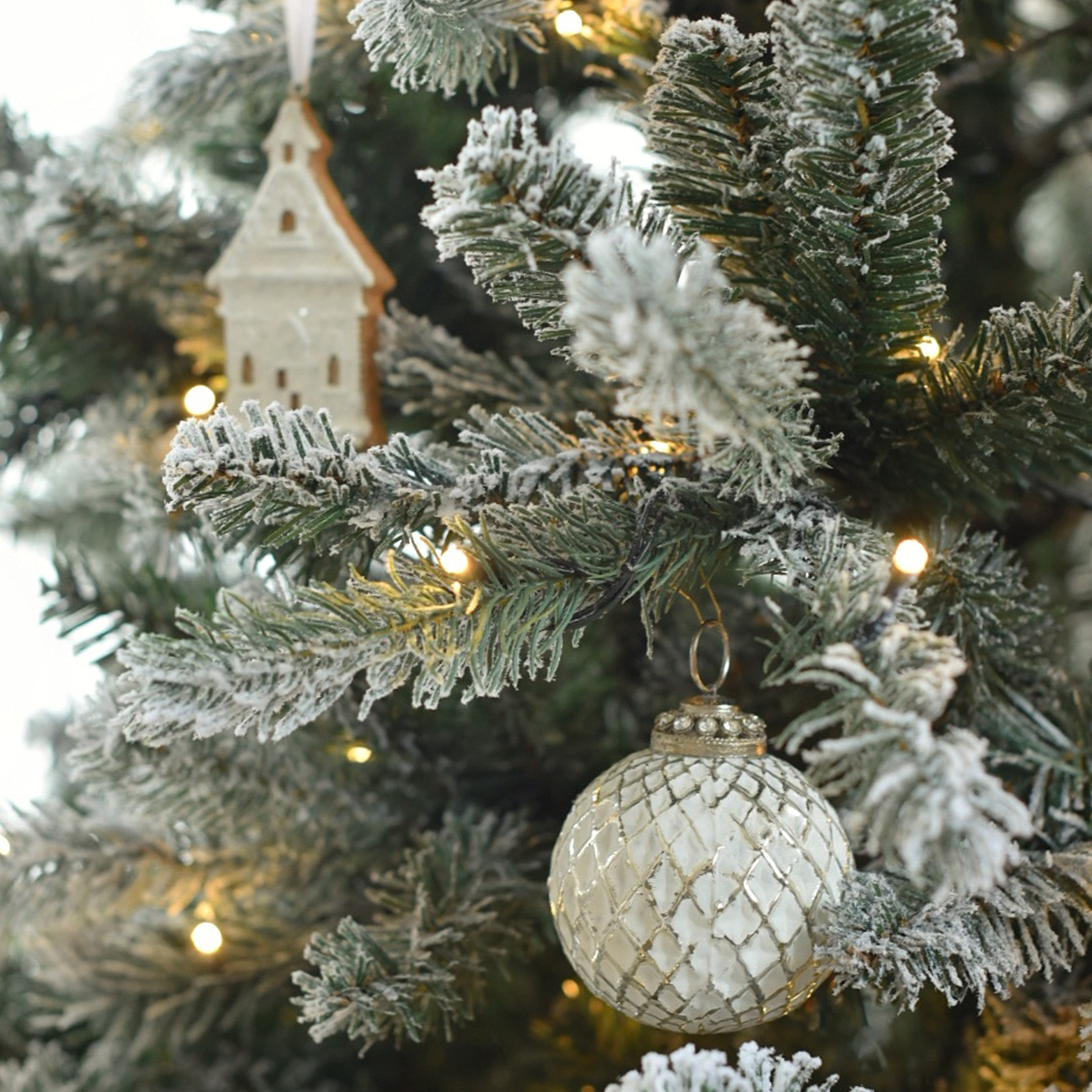 Weiße Glas-Weihnachtskugel mit Rautenmuster an schneebedecktem Tannenbaum mit LED-Lichtern und Holzdeko
