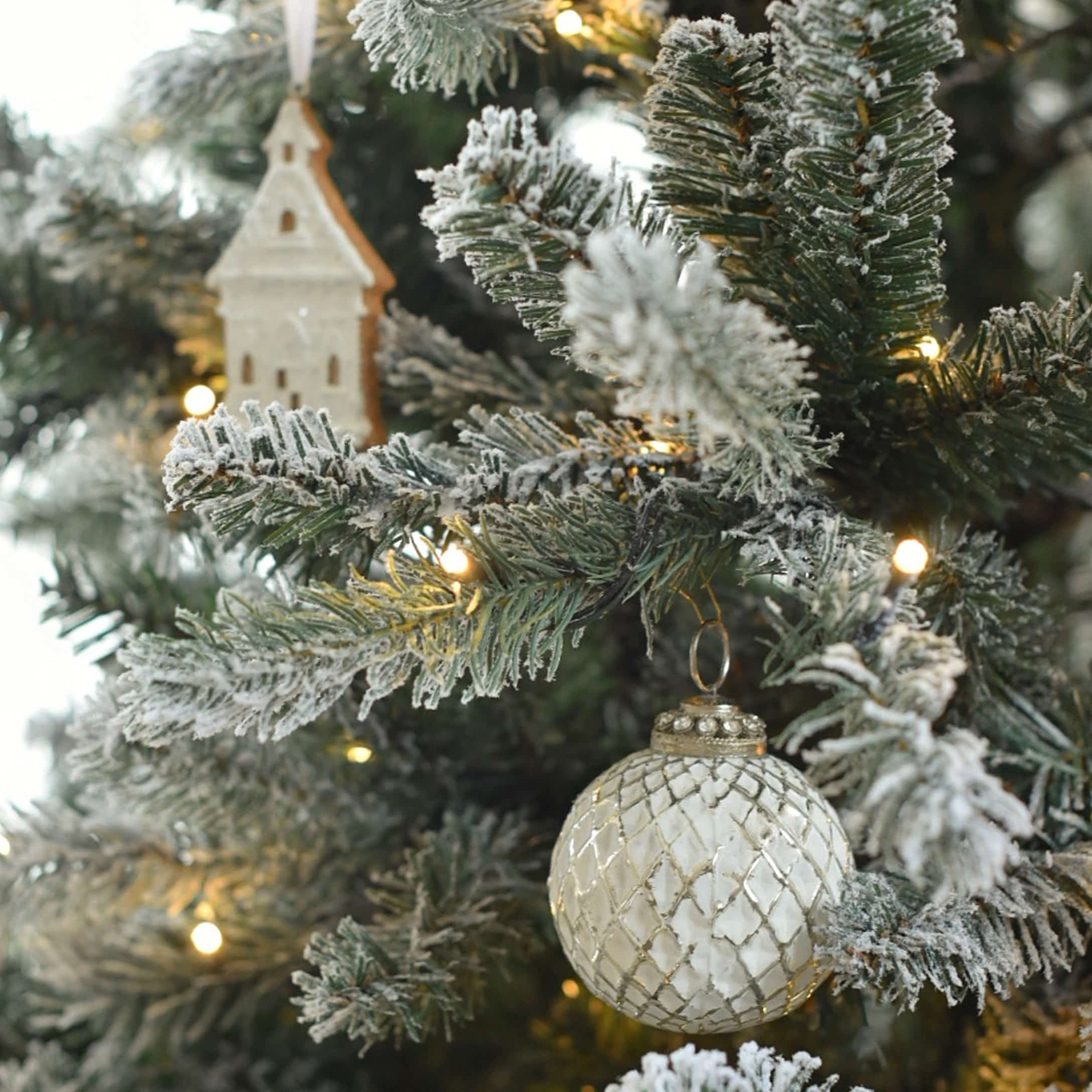 Weiße Glas-Weihnachtskugel mit Rautenmuster an schneebedecktem Tannenbaum mit LED-Lichtern und Holzdeko