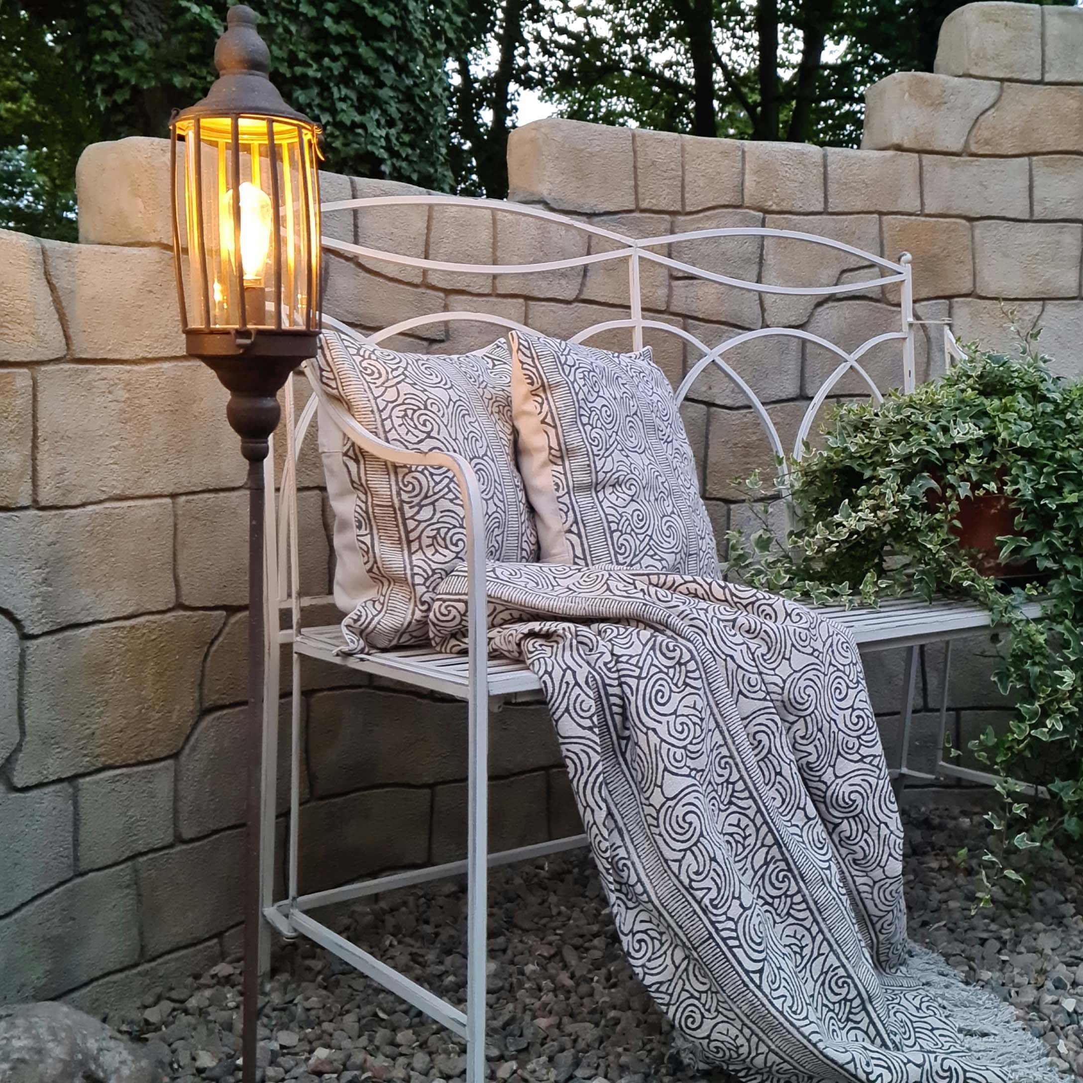 Weiße Metall-Gartenbank mit dekorativen Kissen und gemustertem Plaid, beleuchtete Stehlampe daneben